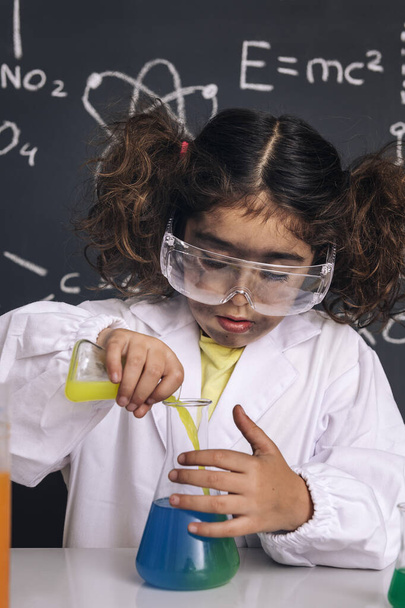 scientist kid with goggles and gloves in lab coat mixing chemical liquids in flasks, blackboard background with science formulas, explosion in the laboratory, back to school concept, vertical photo - Photo, image