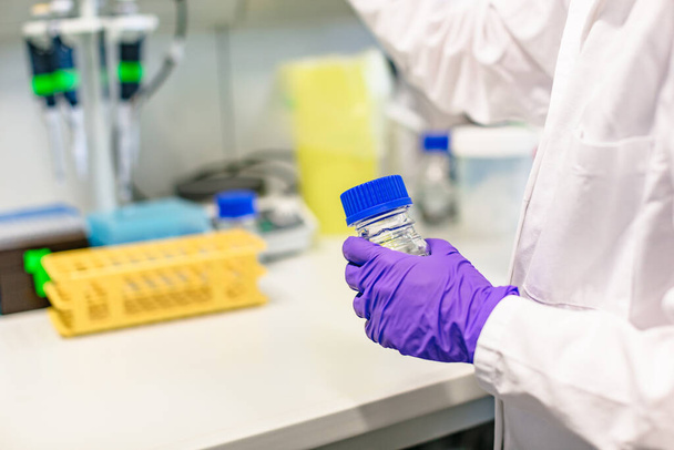 Female hand in blue gloves in bathrobe in laboratory holds bottle - Fotoğraf, Görsel