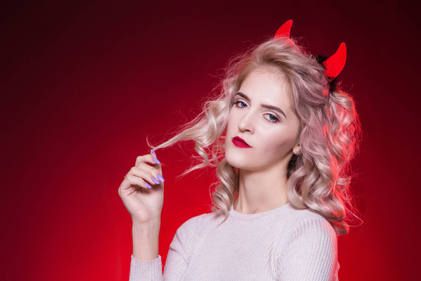 To twist a curl of hair on a finger. Portrait of a beautiful blonde woman with a cunning look and horns on her head. The dress for the Halloween party. Evil demon girl is the devil. - Foto, Imagen