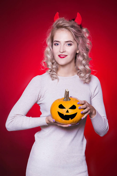 Beautiful blonde woman with a cunning look and horns on her head. The dress for the Halloween party. Holds a pumpkin and smiles. Red Background - Foto, afbeelding
