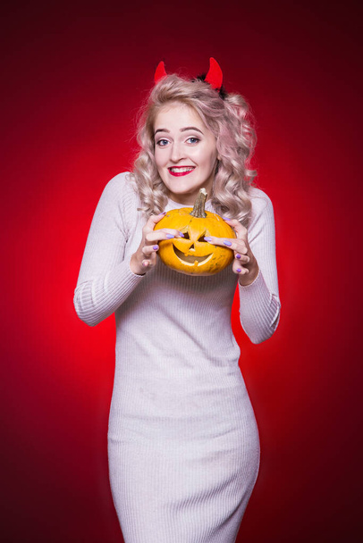 Beautiful blonde woman with a cunning look and horns on her head. The dress for the Halloween party. Holds a pumpkin and smiles. An embarrassed, awkward look. Red Background - Фото, изображение