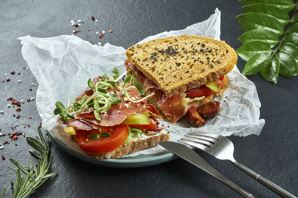 Trendy street snack. Tasty sandwich with jamon, tomatoes, cucumber and microgreen on craft paper on a black background. Food flat lay, copy space. - Foto, Imagem