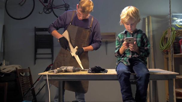 Junge sitzt mit Telefon, Meister arbeitet in Werkstatt mit Holz - Foto, Bild