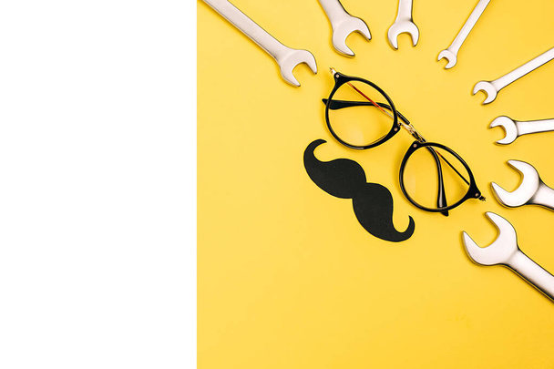 Fathers Day mockup with eyeglasses, combination wrenches and stylish black paper photo booth props moustaches on yellow and white background with copy space. Flat lay, top view, minimal composition - Foto, Imagem