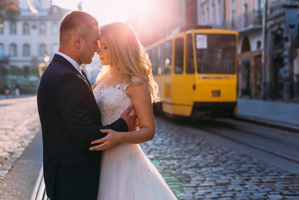 Dantelli beyaz elbiseli güzel bir gelin. Takım elbiseli damat geline sarılıyor. Kare ve tramvayın arka planında yeni evlilerin profili. - Fotoğraf, Görsel