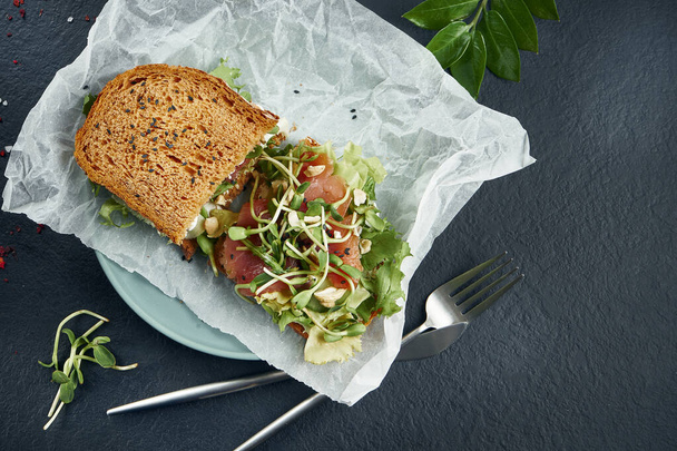 Trendy street snack. Tasty sandwich toast with salmon and microgreen on craft paper on a black background. Top view. Flay lay food with copy space. Seafood - Foto, Imagem