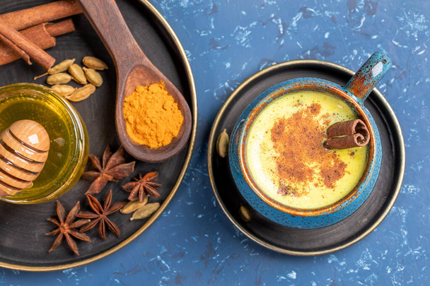 Top view of cup of golden turmeric milk and plate with ingredients: curcuma powder, cinnamon, anise, honey on blue. Traditional Indian ayurvedic turmeric milk is healthy drink for improve immunity. - Фото, изображение