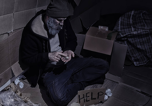 Een dakloze man met een baard zit op dozen op straat en vraagt om hulp. Noodzaak van een dakloze vraagt om geld voor voedsel en 's nachts. - Foto, afbeelding