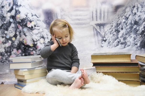 Cute little girl playing with a smartphone sitting with legs crossed surounded by books - Фото, зображення