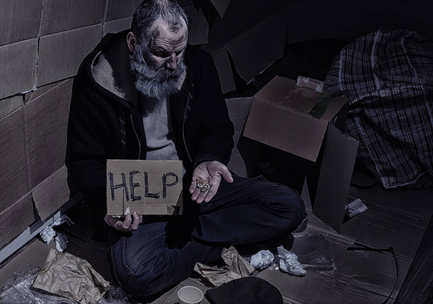 Un senzatetto barbuto si siede su delle scatole per strada e chiede aiuto. Bisogno di un senzatetto chiede soldi per il cibo e durante la notte. - Foto, immagini