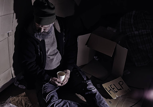 A homeless bearded man sits on boxes on street and asks for help. Need a homeless person asks for money for food and overnight. - Zdjęcie, obraz