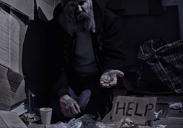Un senzatetto barbuto si siede su delle scatole per strada e chiede aiuto. Bisogno di un senzatetto chiede soldi per il cibo e durante la notte. - Foto, immagini