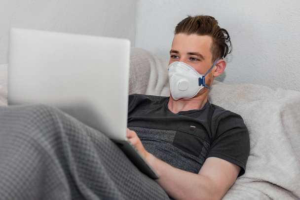 Young an σε μια ιατρική μάσκα, βρίσκεται σε ένα κρεβάτι με ένα φορητό υπολογιστή, εργάζεται από το σπίτι κατά τη διάρκεια της καραντίνας κατά τη στιγμή της επιδημίας coronavirus, covid-19. Φορώντας γκρι μπλουζάκι, καλυμμένο με γκρι καρό - Φωτογραφία, εικόνα