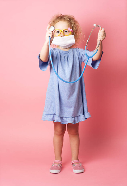 Children's medicine: a little girl in a blue dress and a medical mask holds a stethoscope in her hands. Portrait on a pink background. - Foto, afbeelding
