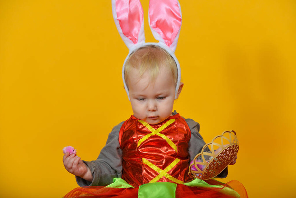 Ένα μικρό παιδί, ένα κορίτσι κλόουν με αυτιά κουνελιού κρατά πασχαλινά αυγά στα χέρια της - Φωτογραφία, εικόνα