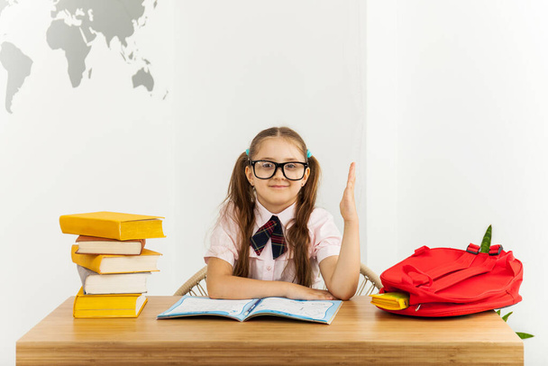 Feliz niña estudiante con gafas con cuaderno abierto tira de una mano con en el escritorio en la sala de clases. Concepto educativo
 - Foto, Imagen