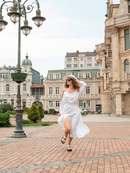 Girl female in a white long dress, with a backpack, holding hat walking near the clock. Back view of attractive and happy girl in old cozy streets of Europe. Pretty sexy fashion model in cityscape. Lifestyle, travel, vacation, tourism - Фото, изображение