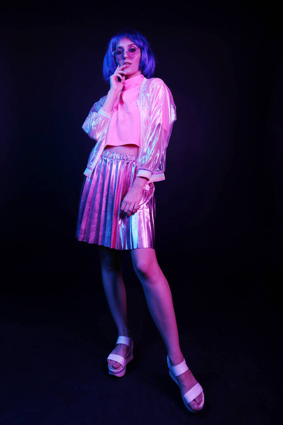 Fashionable young woman in neon light in full growth in a nightclub. The girl is wearing a wig, glasses and a shiny, halo jacket and skirt. - Photo, image