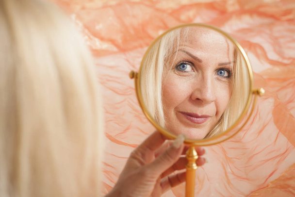 Reflet du visage d'une femme blonde d'âge moyen dans un miroir de maquillage rond sur un fond de tissu orange
. - Photo, image