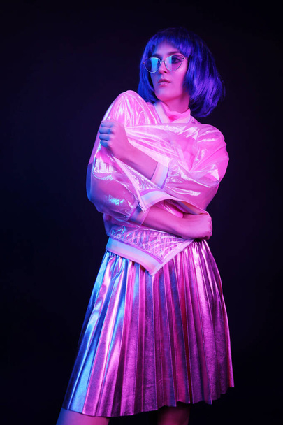 Fashionable young woman in neon light at a nightclub. The girl is wearing a wig, glasses and a shiny, halo jacket and skirt. - Foto, Bild
