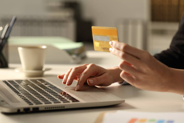 Primer plano de las manos de la mujer ejecutiva con tarjeta de crédito un ordenador portátil que paga en línea por la noche en la oficina
 - Foto, Imagen
