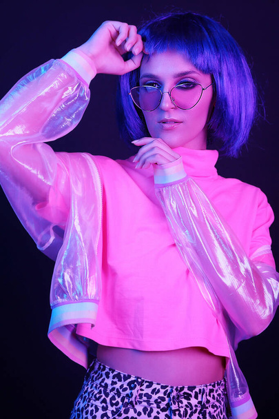 Fashionable young woman in a neon light dancing in a nightclub. The girl is wearing a wig, glasses and a shiny, halo jacket. Copy space. - Photo, image