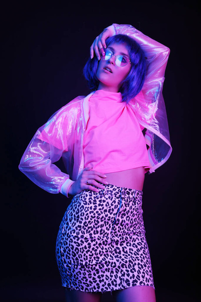 Fashionable young woman in a neon light dancing in a nightclub. The girl is wearing a wig, glasses and a shiny, halo jacket, leopard skirt. - Valokuva, kuva