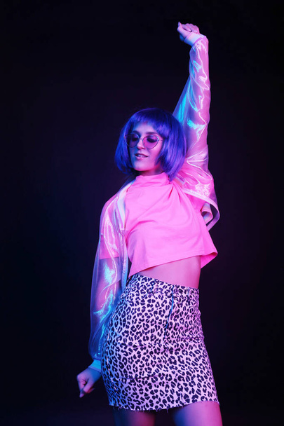 Fashionable young woman in a neon light dancing in a nightclub. The girl is wearing a wig, glasses and a shiny, halo jacket, leopard skirt. - Foto, afbeelding