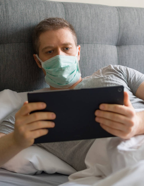 ベッドのタブレットPCを使用して医療用マスクを持つ病気の男。ホーム隔離の概念. - 写真・画像