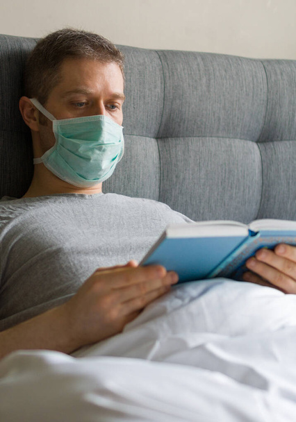 Yatakta maskeli hasta bir adam kitap okuyor. Ev karantina konsepti. - Fotoğraf, Görsel