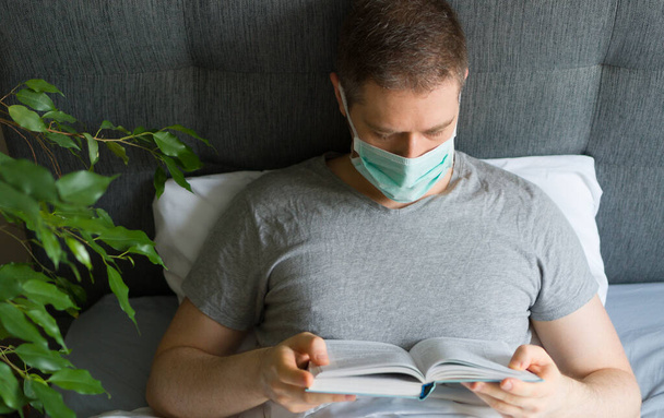 Sick man with medical mask reading book in bed. Home quarantine concept. - Φωτογραφία, εικόνα