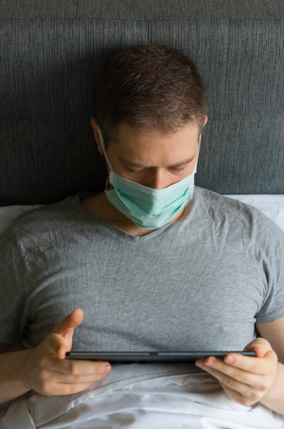 Hombre enfermo con máscara médica usando tableta pc en la cama. Concepto de cuarentena doméstica
. - Foto, imagen