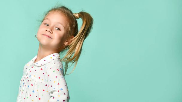 Portrait of a little blond child in a heart-shaped print shirt. She smiles, fools around, has fun against the backdrop of a mint studio - Photo, image