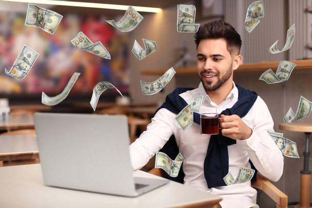 現代のラップトップを持ち、屋内のテーブルでドル紙幣を飛んでいる男。人々はオンラインでお金を稼ぐ - 写真・画像
