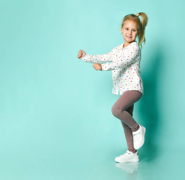 Little blonde girl in shirt with hearts print, checkered pants, white sneakers. Smiling, dancing on blue background. Full length - Photo, Image