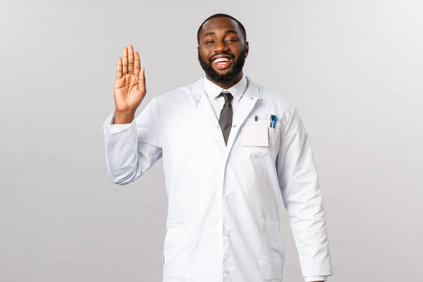 Дружелюбный африканско-американский врач, приглашающий пациентов на осмотр, приветствующий людей с оптимистической радостной улыбкой, машущий рукой, говорящий: никогда не возвращайся в больницу, будь здоров
 - Фото, изображение