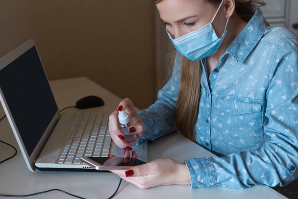 Young woman in face mask disinfecting gadgets surfaces on her workplace - Fotoğraf, Görsel