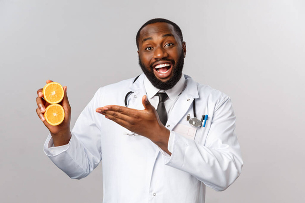 医療、健康的な食事と病気の概念。幸せな陽気なアフリカ系アメリカ人のひげ医者、医師は果物、新鮮なジュース、オレンジと笑顔を示すことの利点を説明します - 写真・画像