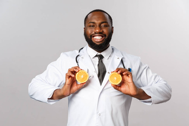 医療、健康的な食事と病気の概念。面白いハンサムなアフリカ系アメリカ人男性医師、医師は喜んで笑顔、胸のような果物を保持し、オレンジと愚か者、灰色の背景に立つ - 写真・画像