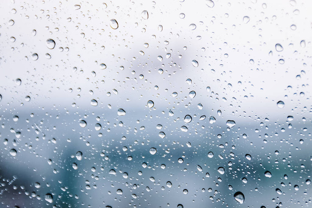 Gotas de lluvia en la superficie del cristal de la ventana húmeda en temporada de lluvias. Fondo abstracto. Patrón natural de gotas de lluvia aisladas de la ciudad borrosa al aire libre en un ambiente nublado
. - Foto, imagen