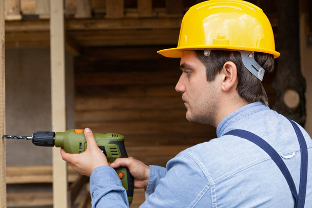 Young handsome carpenter with yellow hard hat and work equipment is holding an electric drill on wooden background - Foto, Imagen