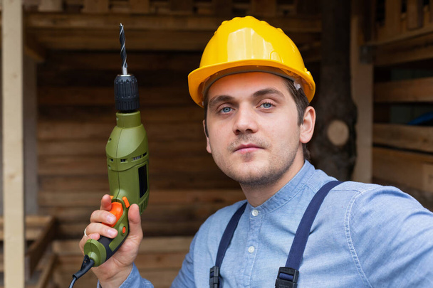 Young handsome carpenter with yellow hard hat and work equipment is holding an electric drill on wooden background - Фото, изображение