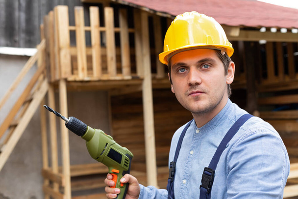 Joven carpintero guapo con sombrero duro amarillo y equipo de trabajo está sosteniendo un taladro eléctrico sobre fondo de madera
 - Foto, Imagen