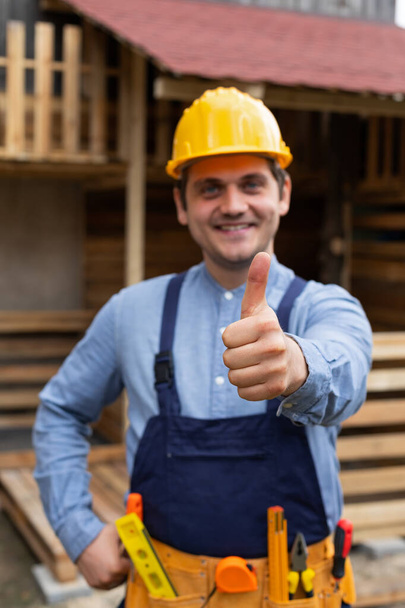 Young carpenter with yellow hard hat and work equipment showing thumbs up in front of wooden background - Fotoğraf, Görsel