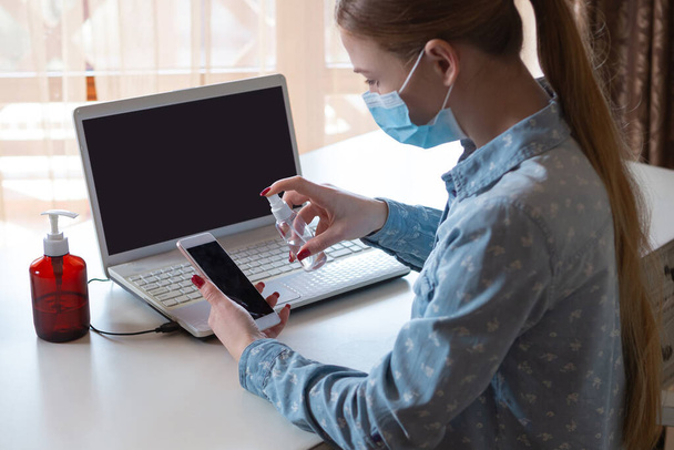 Young woman in face mask disinfecting gadgets surfaces on her workplace - Zdjęcie, obraz