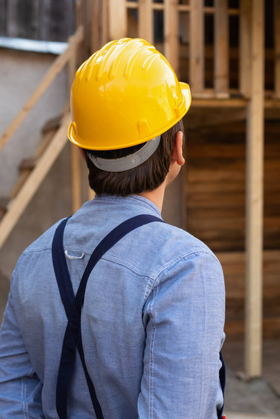 Back view of young carpenter with yellow hard hat and blue shirt outdoor, wooden background - Foto, Imagem