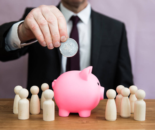 A business concept of a businessman paying into or contributingan employee salary, pension, tax, bonus or incentive scheme by placing money into a piggy bank - Photo, Image