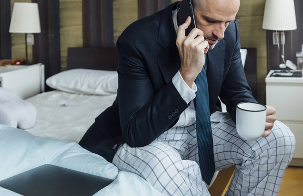 Podnikatel sedí na posteli s chytrým telefonem a kávou - Fotografie, Obrázek