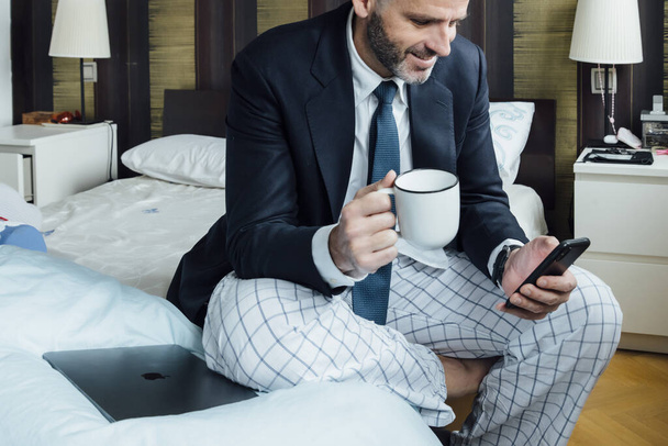 Biznesmen siedzi na łóżku ze smartfonem i kawą - Zdjęcie, obraz