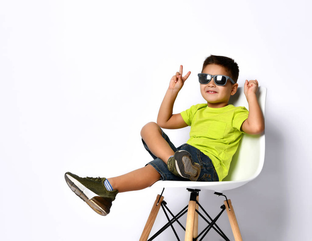 Kid in sunglasses, yellow t-shirt, denim shorts, khaki sneakers. Smiling showing victory sign, sitting on chair. Isolated on white - Φωτογραφία, εικόνα